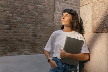 Laptoplu, mutlu, koyu renk saçlı kız güneşli bir şehirde dışarıda bekliyor. Açık havada, dışarıda güneşlenen mutlu bir bayanın portresi..