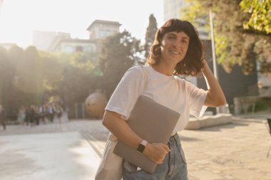 Harika gülüşü ve kısa koyu renk saç stili olan başarılı güzel bayan beyaz tişört ve kot pantolon giyerek yaz kenti parkında dizüstü bilgisayarla kameraya poz veriyor.