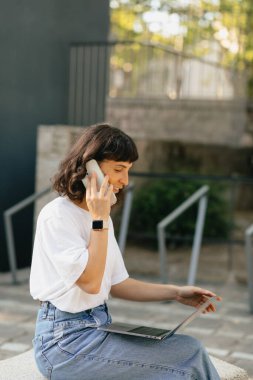 Tarz sahibi beyaz bir kadın dışarıda oturuyor ve akıllı telefondan arkadaşıyla konuşuyor. Kulaklık takan pozitif bir kız günlük kıyafetler giyer. Konsept video bağlantısı, iletişim aygıtları.