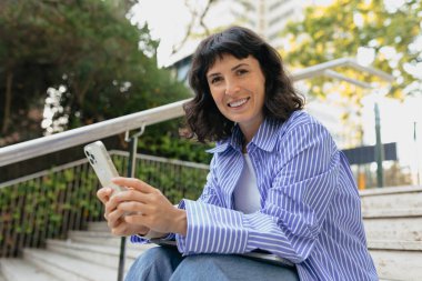 Güzel stil sahibi, güzel Avrupalı bir kadın mutlu gülüşü ve kısa siyah saç stili ile mavi gömlek giyiyor ve akıllı telefon kullanıyor. Akıllı telefonu tutarken kameraya gülümseyen sevimli bayan..