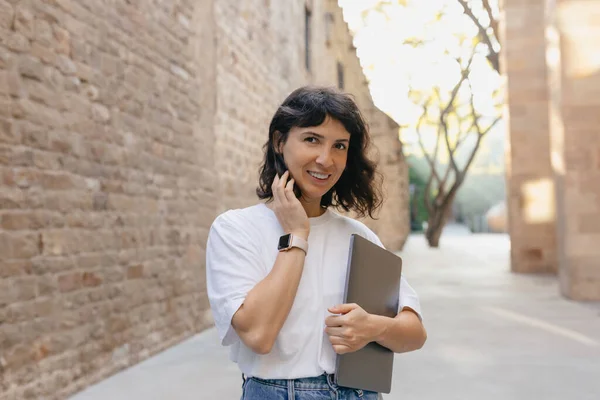Enchanting female freelancer with wonderful smile is smiling to camera. Outdoor photo of refined dark-haired student holding laptop while posing on the street.