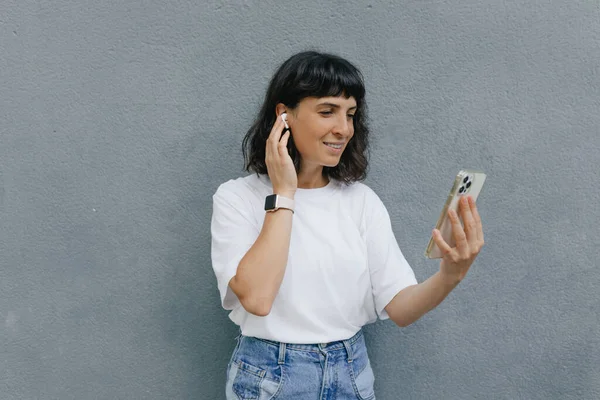 Cute charming woman with dark wavy hairstyle and charming smile wearing white t-shirt and jeans is talking on video call with friend on background grey wall.