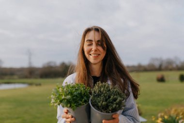 Harika gülüşü ve açık siyah saçları olan sevimli, sevimli bir bayan. Gözleri kapalı ve güneşli bir günde yeşile bürünmüş taze bitkilerle gülümsüyor.