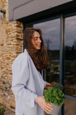 Güzel, genç, esmer Avrupalı bir kadın yaz terasında taze bitkiler tutuyor ve gülümsüyor. Model gömlek ve kot pantolon giyer. Yaşam tarzı konsepti samimi duygular