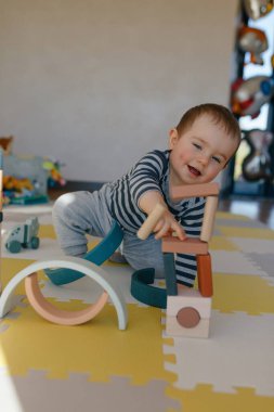 Küçük çocuk ahşap eğitici oyuncaklarla oynuyor. Süslü püslü gömlekli çocuk evinde yerde oturuyor ve ahşap elementlerle kale inşa ediyor. 