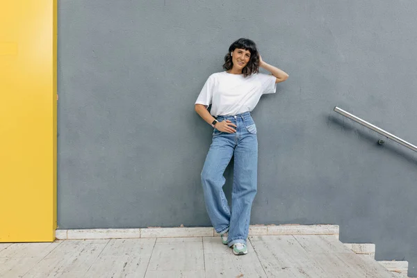 stock image Full-length photo of stylish good-looking woman with short hairstyle wearing white t-shirt and jeans is holding up hand and smiling outdoor over grey wall