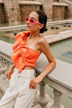 Oldukça çekici, şık saçları olan zarif kızlar parlak bir yaz bluzu giyiyorlar ve parlak gözlüklü pantolonlar şehirde gülümseyerek ve dinlenerek kameraya poz veriyorlar..