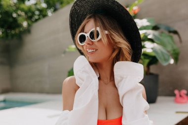 Harika gülüşlü, kısa sarı saçlı, siyah şapkalı güzel bir kadının profil portresi. Beyaz gömlek, arka planda havuz ve egzotik bitkilerin üzerinde dinleniyor. 