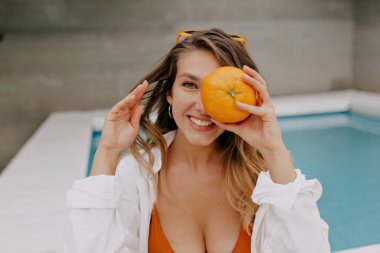 Uzun dalgalı saçlı, turuncu mayo giyen, beyaz gömlekli sevimli kız kameraya gülümsüyor, yüzünü turuncu ile kaplıyor ve havuz kenarında dinleniyor. 