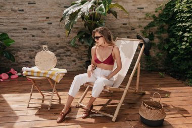 Güneş ışığında egzotik bir tatil köyünde üstsüz, gözlüklü ve beyaz pantolonlu zarif güzel bir kadının tam boy fotoğrafı. Açık havada, turuncu elbiseli harika bir kızın deniz kenarında kumlarda serinlerken görüntüsü.. 