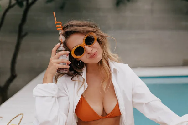 オレンジの水着と白のシャツを着て波状の髪を持つかわいい可愛い女性とスタイリッシュなメガネは プールを背景にカメラにポーズをとって夏のドリンクを開催しています — ストック写真