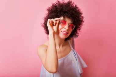 Kıvırcık saçlı, yuvarlak gözlüklü ve yaz tişörtlü sevimli bir kadının portresini yakın çekimde pembe arka planda kameraya çektiğimi varsayıyorum. 