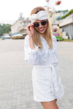 Romantik, çekici, zayıf, sarı saçlı, beyaz yaz kıyafetleri giyen, gözlüklü genç bayan gözlüklerine dokunuyor ve şehirde dışarıda vakit geçiriyor. 