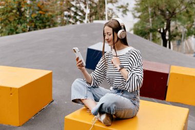 Açık renk saçlı, çizgili gömlekli ve kot pantolonlu güzel bir kızın uzun metrajlı fotoğrafı akıllı telefon kullanıyor ve yaz günü dışarıda müzik dinliyor..