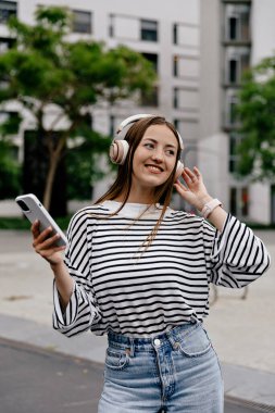 Harika, oldukça güzel saçlı, yüzgeçli tişörtlü, kot pantolonlu, kulaklıklı yaz günü dışarıda akıllı telefonuyla bekleyen bir kız.. 