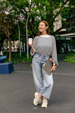 Gülümseyen güzel bir kadının uzun boylu fotoğrafı elinde bir fincan kahve ve dizüstü bilgisayarla şehirde mutlu bir gülümsemeyle yeşil ağaçlara doğru yürüyor. 