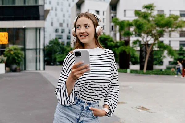 Pretty good-looking girl with smartphone in hands and wearing headphones is enjoying music and looking aside with happy smile