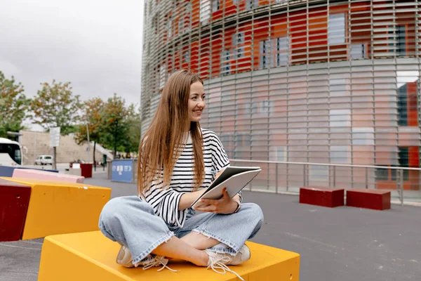 Outside photo of lovely cute girl with loose hair wearing striped shirt and jeans is sitting with notebooks. Smiling european blonde in casual clothes. Concept of outdoors work time.
