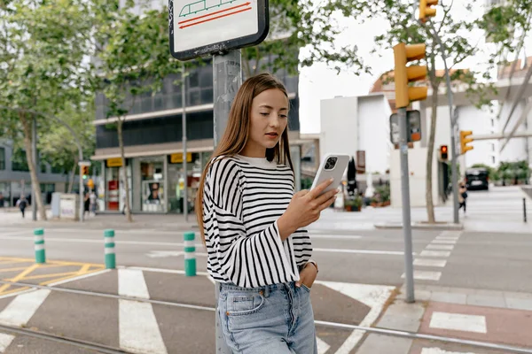 Cute young caucasian girl wears jeans and striped shirt stands outdoor, uses smartphone. Brunette hair woman texting messages in chat. Concept social life, lifestyle.