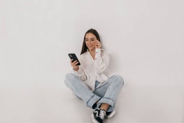 Gömlekli ve kotlu modern bir kızın gözlüklerine dokunduğu ve akıllı telefondan büyüleyici bir fotoğraf.