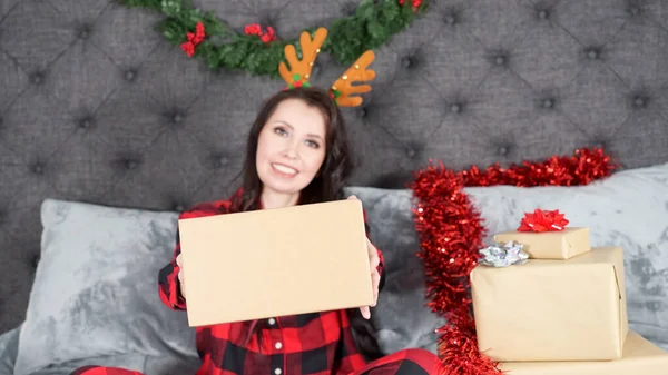 Happy young woman wearing red flannel pajama with holiday deer headband and holding gift box with space for text sitting on bed with Christmas decorations, presents, copy space. Selective focus