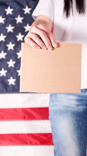 Unrecognizable woman showing empty cardboard banner. Hands holding blank poster from a cardboard box. Space for your slogan, logo or advertisement. Banner design concept. American flag on background.