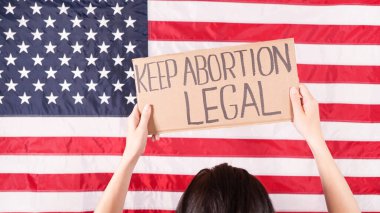 Woman is holding an empty cardboard with Space for Text sign US flag on background. Protest against anti abortion law. Women's strike. Womens rights freedom. Against racism racial discrimination.