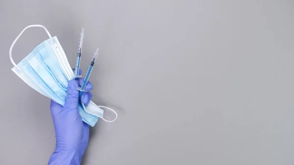 Hand in blue gloves of doctor or nurse holding syringe with liquid vaccine and protective blue face mask over grey background with copy space. New vaccine. Unknown vaccine. Medical gloves