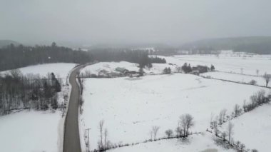 Aerial drone view of beautiful winter scenery in the mountains with pine trees covered with snow and farm at distance. Dark skies and heavy snow falling. Cinematic shot. Winter traveling