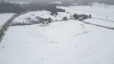 Aerial drone view of beautiful winter scenery in the mountains with pine trees covered with snow and farm at distance. Dark skies and heavy snow falling. Cinematic shot. Winter traveling