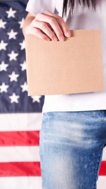 Young woman holds empty cardboard with Space for Text sign against American flag on background. Girl protesting anti-abortion laws. Feminist power. Womens rights freedom