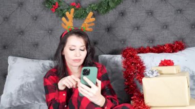 Kırmızı ekose pijamalı genç bir kadın boynuzlu bir saç bandı takıyor Noel süslemeli bir odada hediyelerle sıkılmış bir şekilde telefona sarılıyor.