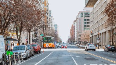Otobüsler, arabalar, yüksek binalar, mavi gökyüzü, yeşil ağaçlar ve bir minibüsle dolu bir şehir manzarası. Güneş ışığı parlak bir atmosfer yaratır. Ön planda alçak açılı bir otobüs durağı görünür.