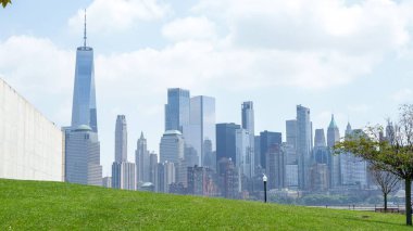 Güneşli bir günde Brooklyn Köprüsü Parkı 'ndan Manhattan' ın nefes kesici manzarası. Açık mavi gökyüzü, beyaz bulutlar, parlayan güneş, yeşil çimenler. Gökdelenler, arka planda apartmanlar.