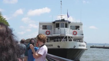 Beyaz ve mavi bir feribot şehrin silueti olan bir rıhtıma yanaştı. Tekne güvertede insanlarla birlikte büyük bir Amerikan bayrağı sallıyor. Şehir manzarası yüksek binaları içeriyor.