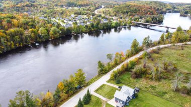 Görüntü, sonbahar mevsiminde suyun yakınında bulunan pitoresk küçük bir köy gösteriyor. Renkli ağaçlar çarpıcı bir zemin oluştururken, huzurlu bir yol büyüleyici topluluğa açılıyor..