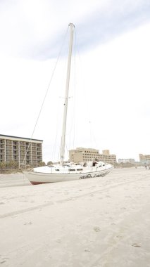 A beached yacht rests on the sandy shore after a hurricane. The damaged vessel sits askew under a gloomy sky as waves crash against the coastline, creating a scene of destruction and despair. clipart