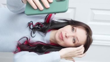 Güzel gülüşü ve dalgalı kahverengi saçları olan bir kadın video görüşmesinde selfie çekiyor, mavi kazak giyiyor, beyaz arka plana karşı, sanal iletişimi ve modern teknolojiyi sembolize ediyor..