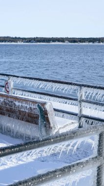 Işıldayan buz saçaklarıyla süslenmiş güzel ahşap bir bank, deniz kenarında zarif bir şekilde oturur. Yumuşak, beyaz kar ve ışıl ışıl buzla çevrili, huzurlu bir kış manzarası yaratıyor, sessiz bir yansıma yaratıyor..