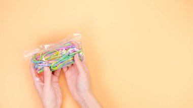 Şeftali arka planında içi renkli lastiklerle dolu şeffaf bir poşet tutan bir el. Lastik bantlar pembe, mavi, yeşil ve sarı olmak üzere çeşitli renklerdedir..