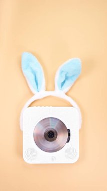 The image features a vintage white portable CD player, elegantly designed, adorned with a playful pair of blue bunny ears headband. Set against a soft peach color background, it evokes nostalgia. clipart