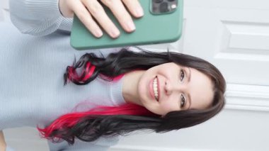 Güzel gülüşü ve dalgalı kahverengi saçları olan bir kadın video görüşmesinde selfie çekiyor, mavi kazak giyiyor, beyaz arka plana karşı, sanal iletişimi ve modern teknolojiyi sembolize ediyor..