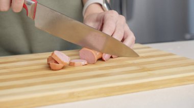 Mutfak ortamında farklı malzemelerle ahşap kesim tahtasıyla sosis kesen bir kadının yakın çekimi.