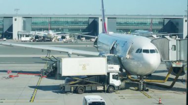 Montreal, QC Canada - 09.12.2024: Havaalanı kapısına bir uçak park edilmiştir. Bir kargo kamyonu bagajları boşaltıyor. Terminal binası arka planda görünüyor. Açık bir gün..
