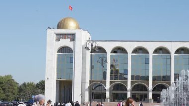 Bişkek, Kırgızistan - 09.15.2024: Altın kubbeli ve büyük cam pencereli modern bir bina. İnsanlar güneşli bir günün tadını çıkarmak için toplandılar. Sahne şehir yaşamını ve mimarisini yakalıyor..