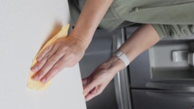 Bir kişi modern bir mutfakta mutfak tezgahını temizliyor. El, beyaz tezgahın üzerinde sarı bir bez olan masayı temizliyor. Mutfak iyi aydınlatılmış ve düzenlenmiş.