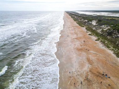 Sahil geniş ve kumlu ve okyanusa doğru yumuşak bir yamaç var. Su berrak ve mavi-yeşil. Gökyüzü dağınık bulutlarla mavidir. Kumsalın arkasında deniz yulaflarıyla kaplı kumullar var..