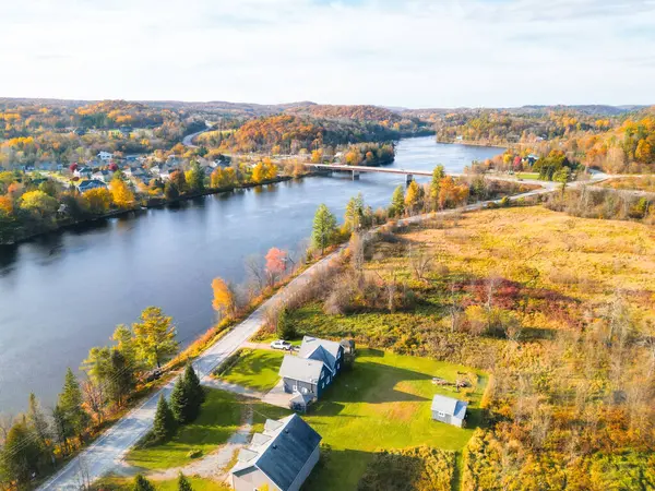 Manzaralı bir kırsal bölgede akan bir nehir, canlı sonbahar ağaçları ve deniz kenarında rahat bir ev sergilenir. Beyaz bulutlu açık mavi gökyüzü huzurlu atmosferi güçlendirir..