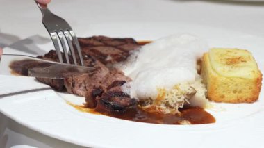 Biftek, pilav ve bir dilim mısırlı ekmeğin yer aldığı yakın plan bir yemek. Bir el çatalı tutar, eti kesmeye hazır..