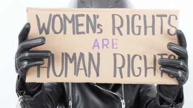 Siyah motosiklet kaskı ve deri eldiven giyen bir kızın elinde Kadın Hakları İnsan Hakları tabelası var. Arkaplan düz beyaz, mesajı vurguluyor.